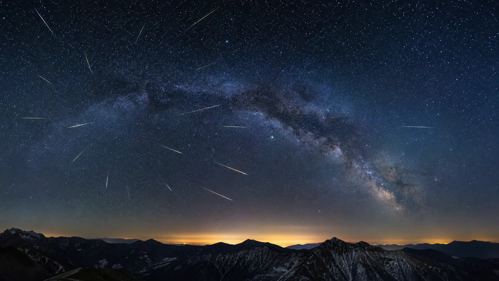 山の稜線の上に広がる満天の星空を、放射状に流れる複数の流星が横切る流星群の夜空