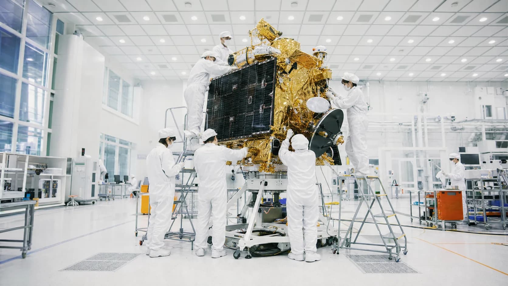 クリーンルームで大型の人工衛星を組み立てる、防塵服を着た複数のエンジニアの写真