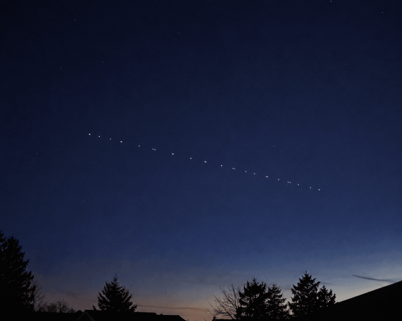 薄明の夜空を、等間隔の光の点が一列に連なって横切るスターリンク衛星。地上には木立のシルエットが見える。